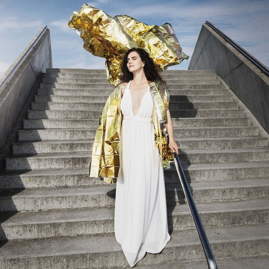Danae Dörken Porträt von Pianistin Danae Dörken auf einer Freitreppe. Weißes Kleid mit goldfarbenem Mantel, moderne Inszenierung bei Tageslicht.
